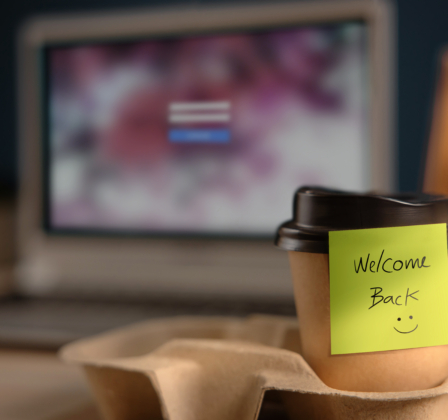 Closeup of a coffee cup in a takeaway holder with a computer screen in the background.